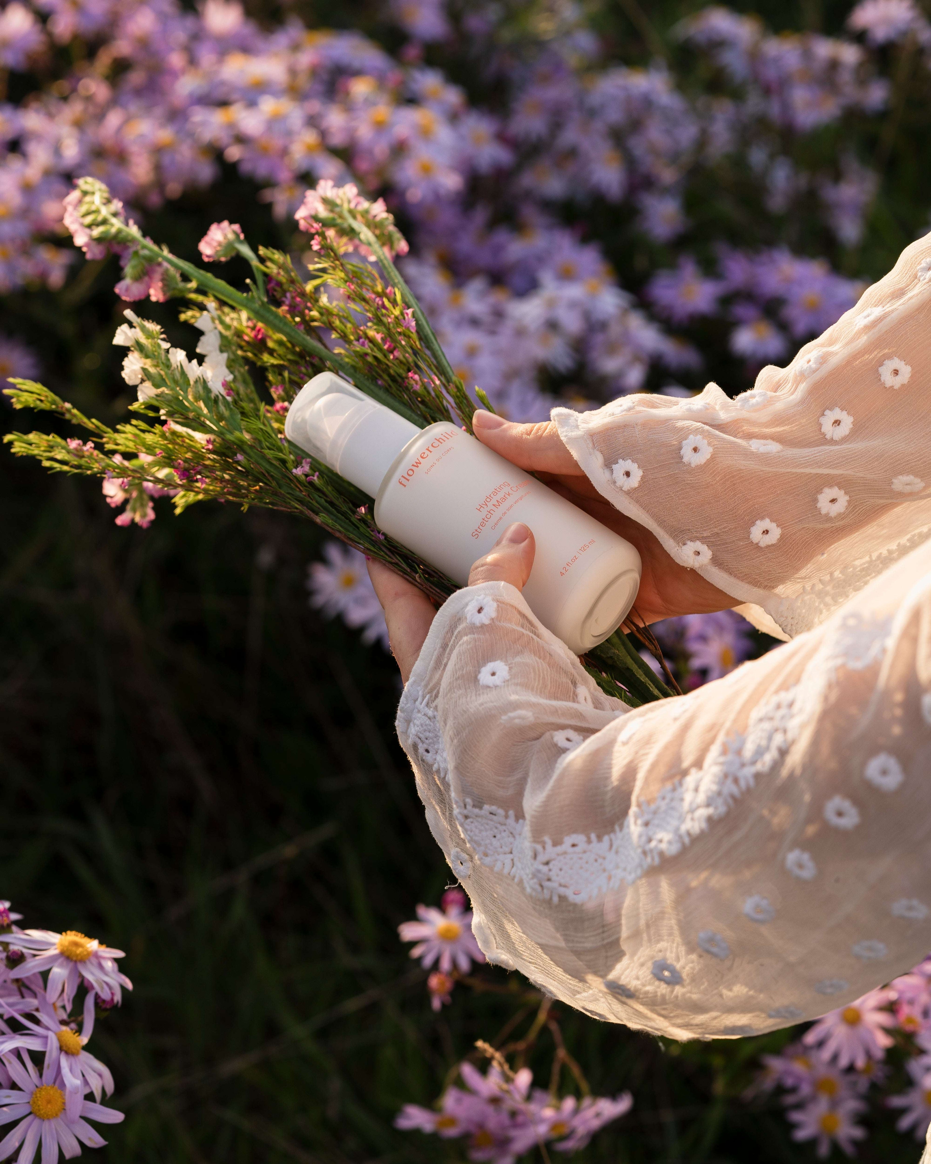 Main féminine tenant la crème anti-vergetures Flowerchild, flacon blanc avec pompe, entouré de fleurs violettes dans un champ naturel.