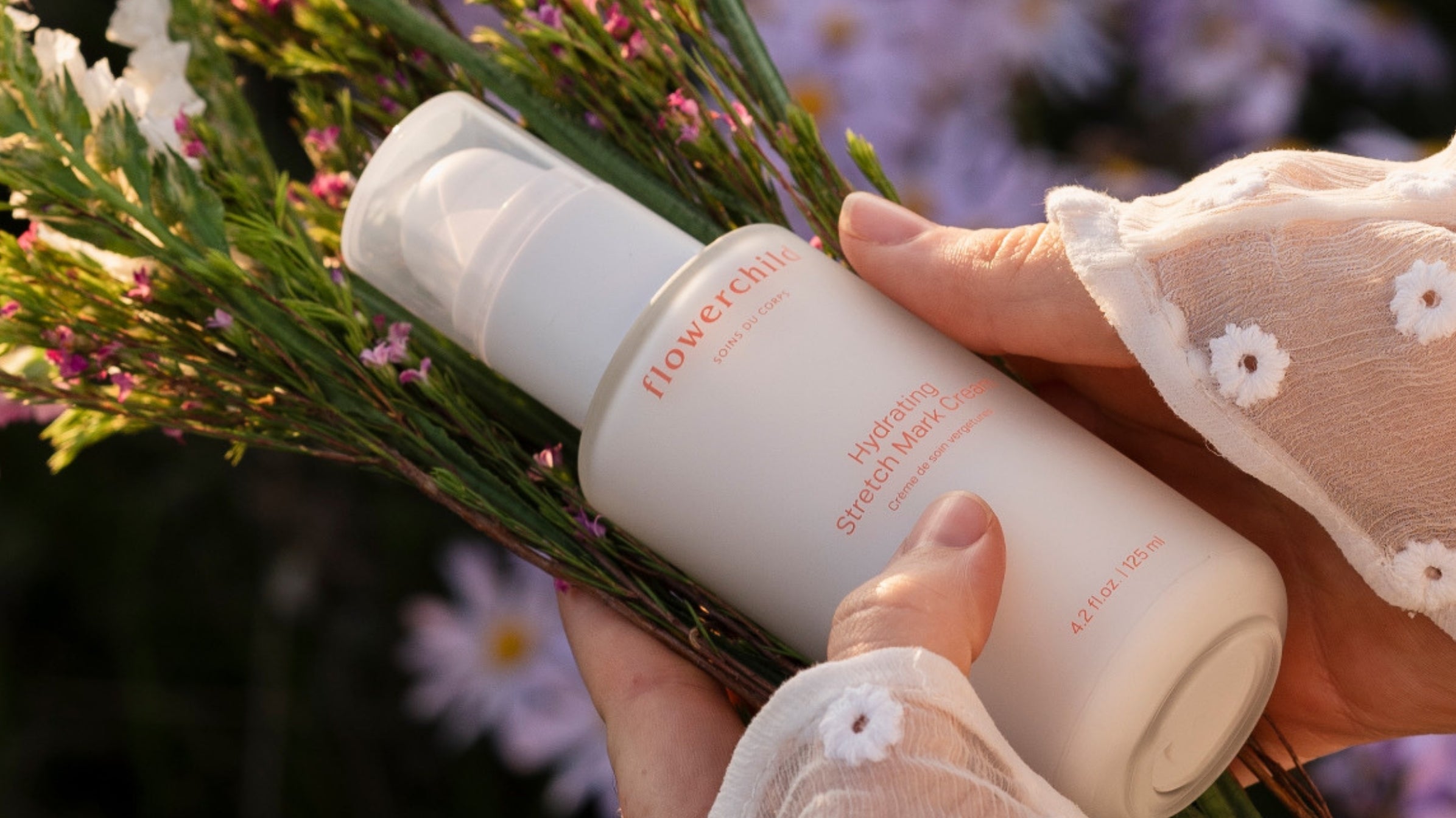 Crème hydratante vergetures Flowerchild tenue dans les mains avec un bouquet de fleurs sauvages, soin naturel pour la maternité.