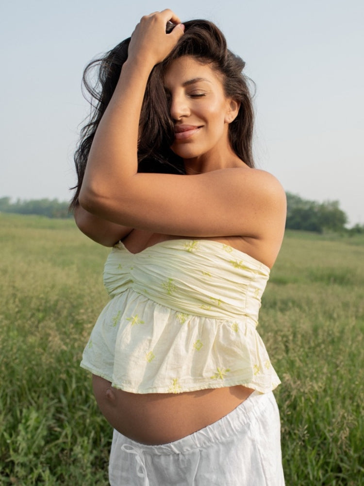 Femme enceinte rayonnante dans un champ en été, portant un haut jaune pâle et un pantalon blanc, capturée en lumière naturelle.