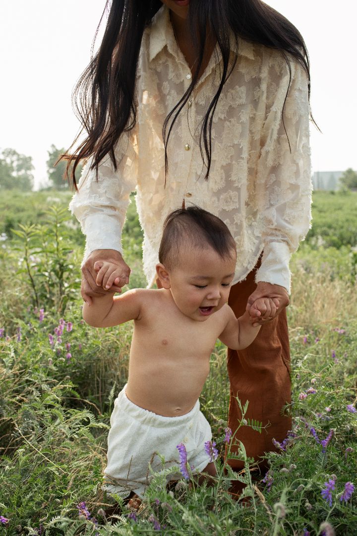 Maman tenant son bébé par les mains dans un champ fleuri, moment complice en plein air.